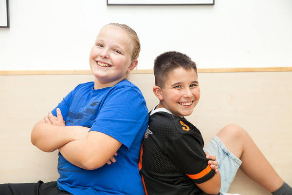 2 Jungs sitzen Rücken an Rücken und grinsen in die Kamera.