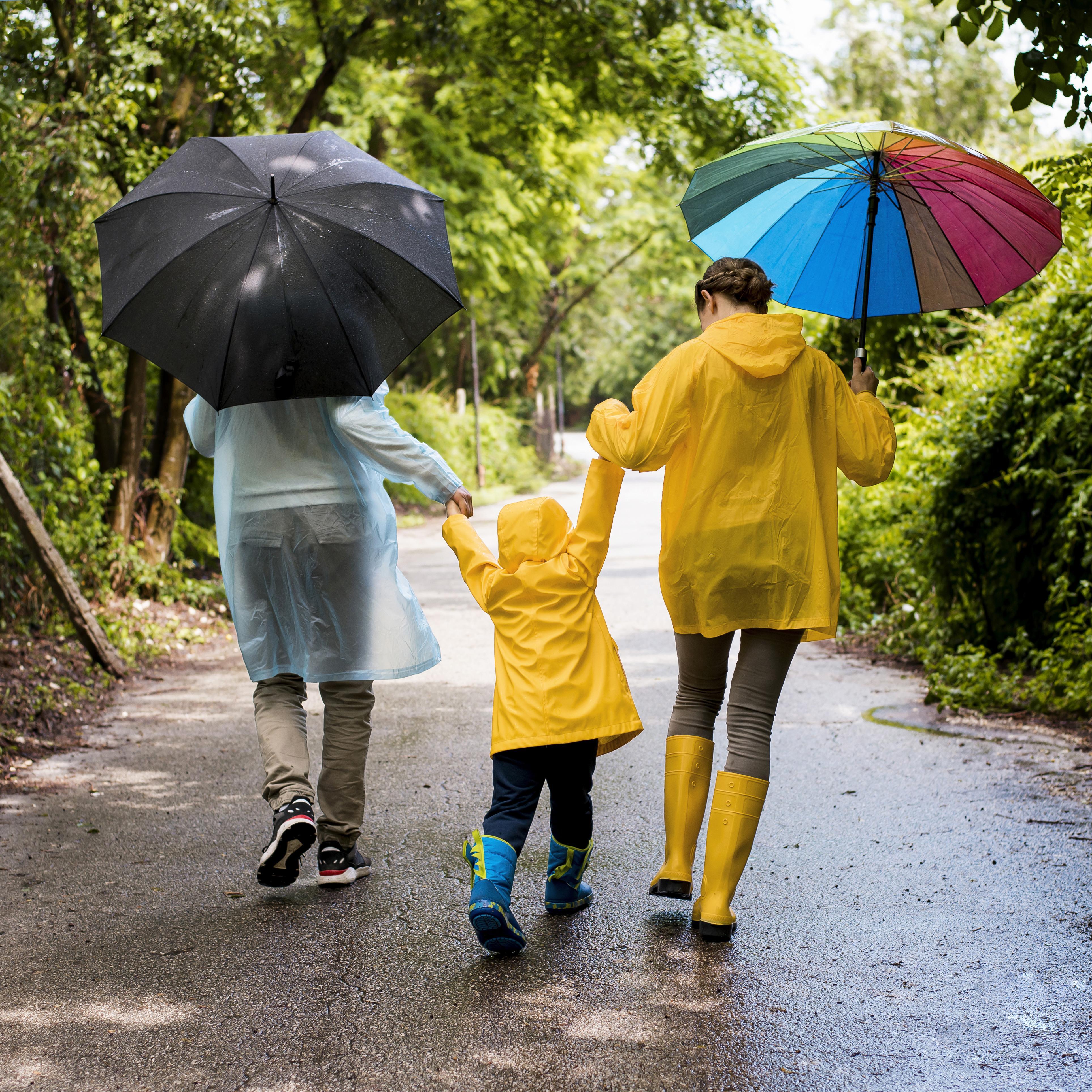 Kind geht mit seinen Eltern im Regen spazieren. Die Eltern halben beide jeweils einen Regenschirm in der Hand. Alle drei tragen eine Regenjacke.
